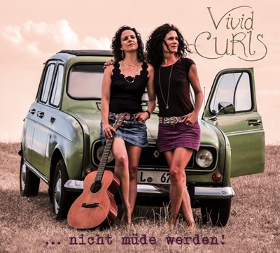 Zwei Frauen der Gruppe Vivid Curls lehnen mit einer Gitarre an der Motorhaube eines Autos, darunter steht &quot;...nicht müde werden!&quot;